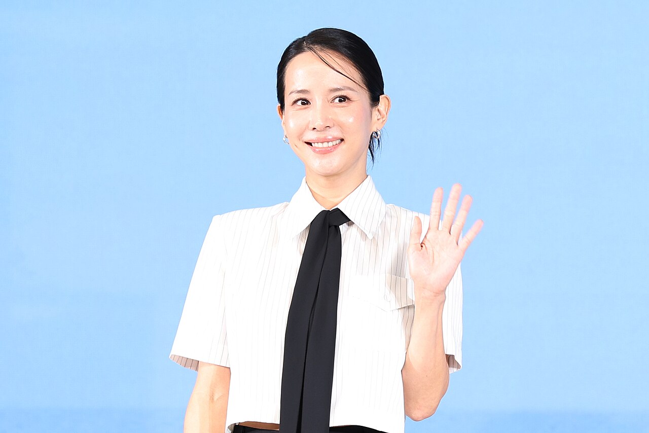 흰 줄무늬 셔츠에 검은 타이를 맨 배우 조여정이 웃으며 손을 흔드는 모습입니다.